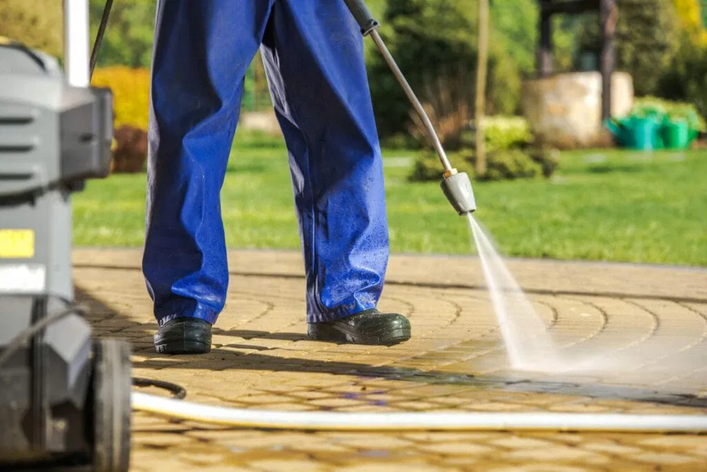 Person in blue pants using a pressure washer for exterior cleaning of outdoor stone tiles in a garden area.